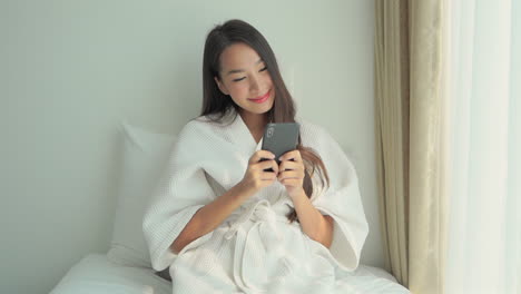 목욕 가운에 침대에 그녀의 전화에 스크롤 웃는 젊은 아시아 아가씨
