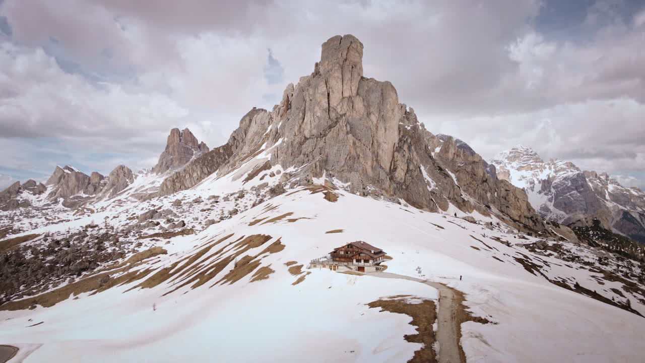 Camera drifts backward from a lone alpine hut, widening to reveal sweeping snow-dusted slopes and the towering Dolomite spires under a dramatic, cloud-filled sky