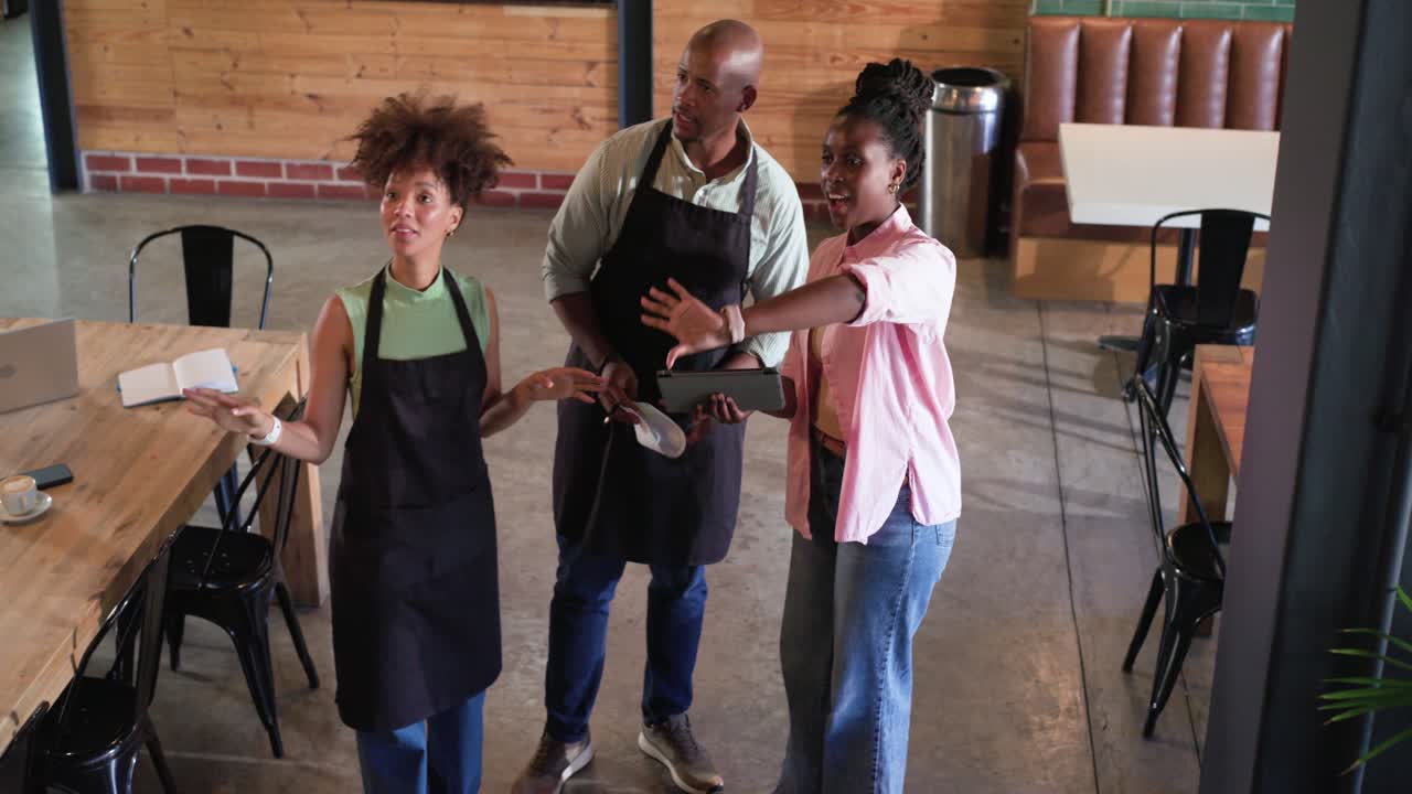 Team afroamericano in un bar, con grembiuli, consulta un tablet e dei documenti al tavolo, mentre il tablet guida la pianificazione