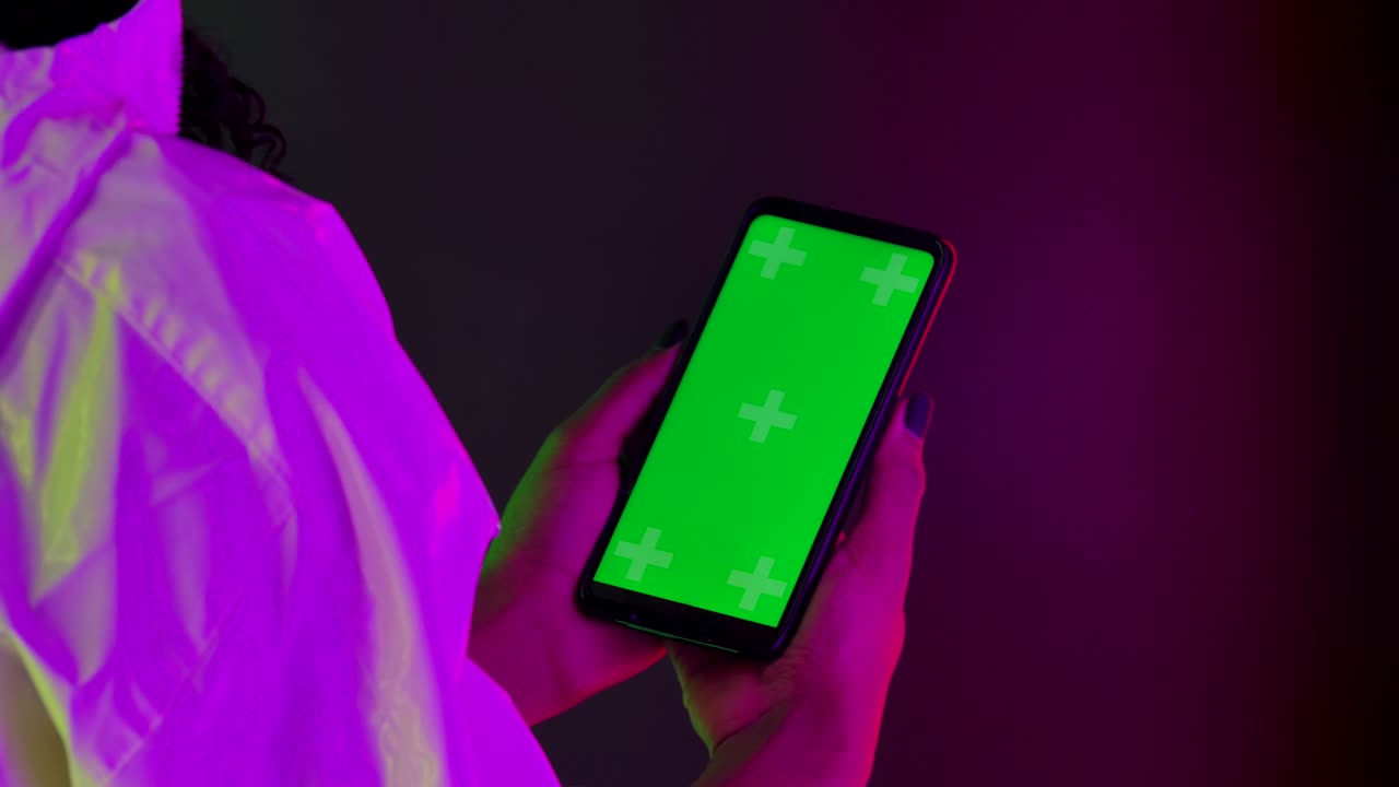 View from behind an African American woman holding a smartphone with a green screen chroma key in her hand. Close up of a phone against a background of bright multicolored neon lights. Slow motion