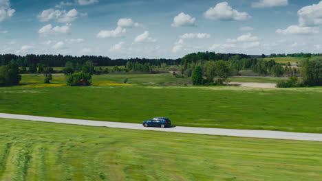 Sideway-drone-shot-of-a-blue-BMW-car-driving-down-a-scenic-road-surrounded-by-lush-green-meadows-on-a-sunny-day
