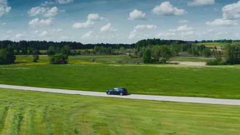 Seitliche-Drohnenaufnahme-Eines-Blauen-BMW-Autos,-Das-An-Einem-Sonnigen-Tag-Eine-Malerische-Straße-Entlang-Fährt,-Umgeben-Von-üppigen-Grünen-Wiesen