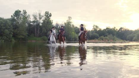 Three-girls-horseback-riding-across-the-river-during-sunset,-handheld-shot