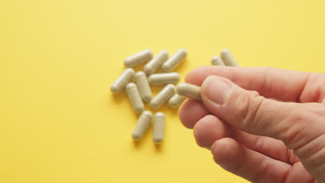 right male hand holding lions mane supplement pill in between fingers with yellow background with pills on table