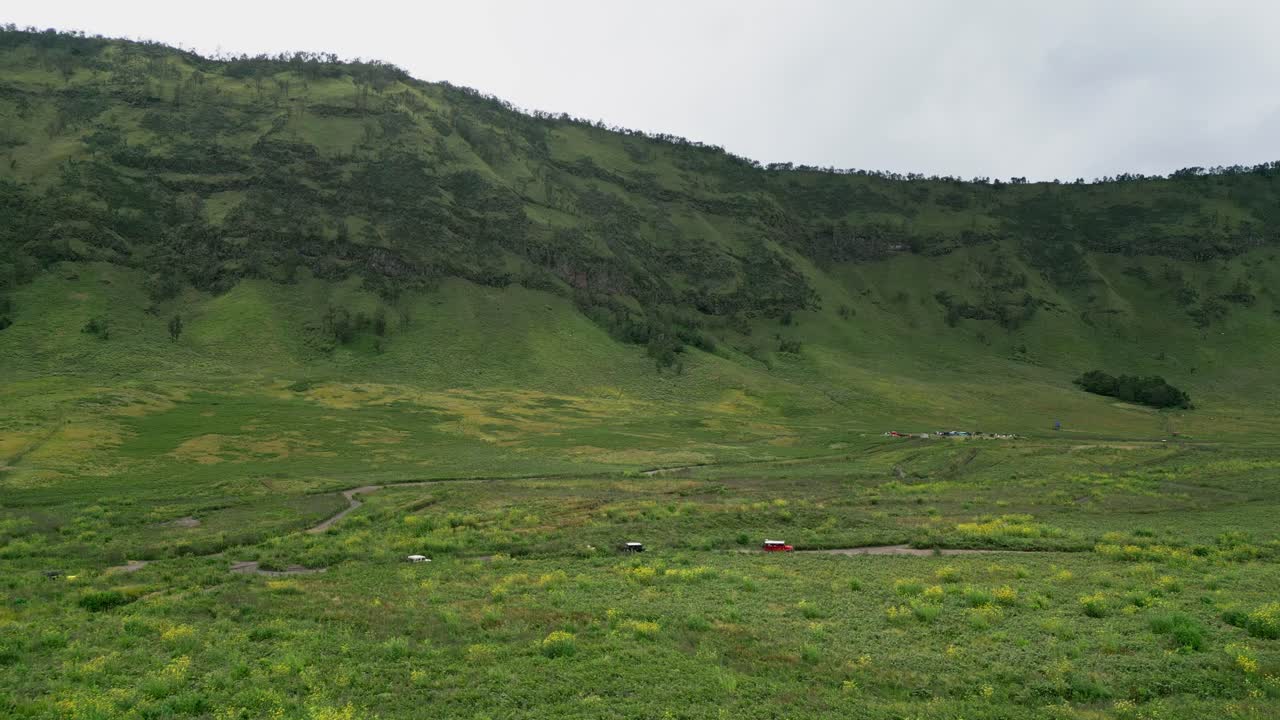 Filmato aereo realizzato con drone che mostra un paesaggio di savana verde con jeep fuoristrada che attraversano una prateria aperta, sullo sfondo ripide scogliere verdi e un ampio terreno montuoso a Giava Est, Indonesia