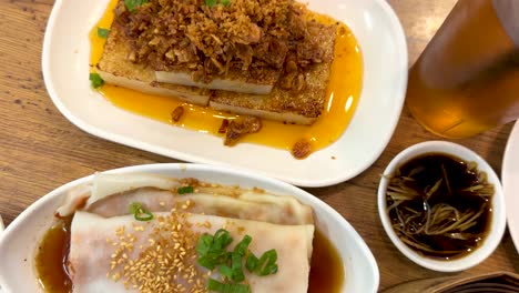 Overhead shots of assorted Singaporean dishes including fried noodles, turnip cake, and rice rolls, with vibrant sauces and tea, under bright indoor lighting
