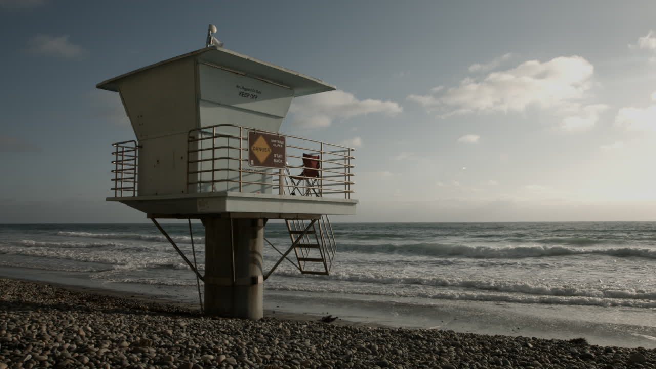 UHD footage captures a peaceful lifeguard tower on the sands of Torrey Pines State Beach near San Diego, bathed in soft golden hour light, evoking a sense of calm, relaxation, and coastal serenity.