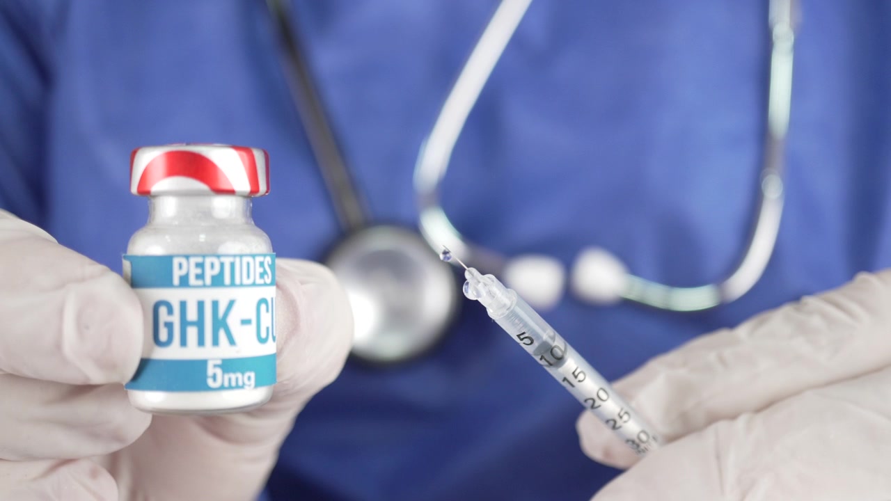 Gloved medical professional holds a GHK CU peptide vial and loaded syringe in front of blue scrubs and stethoscope, useful for content on injections, peptides, or clinical procedures.