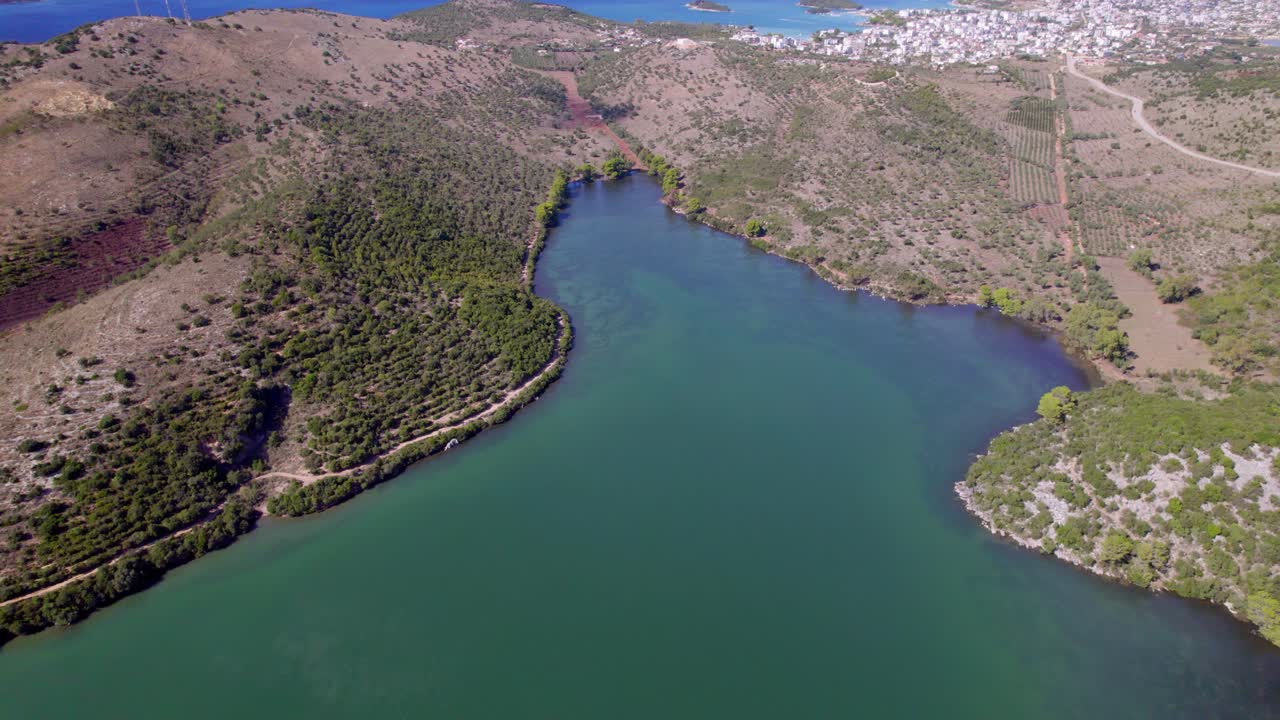 Vista de drone em alta altitude de uma lagoa calma cercada por colinas verdes com uma cidade litorânea e oceano azul ao fundo, movimento suave de panorâmica da câmera