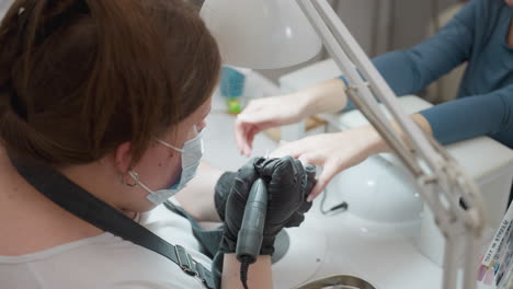top view of nail technician with tied blond hair wearing face mask and apron, intensely focused while filing client nails with electric tool and brush under bright lamp in clean salon setup