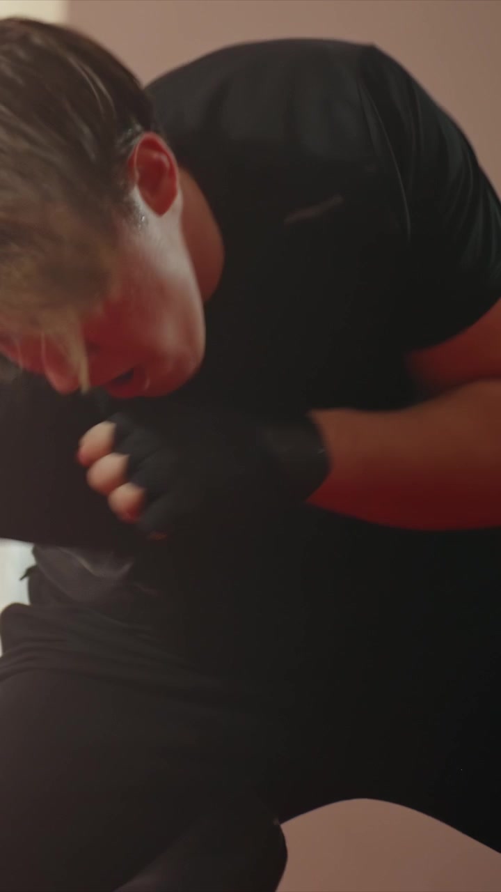 Giovane uomo in maglietta nera pratica la boxe. Le sue azioni includono pugni. L'uomo in seguito si sdraia sulla schiena per fare ripetuti sollevamenti della parte superiore del corpo su un tappetino rosso, mostrando capelli castani corti.