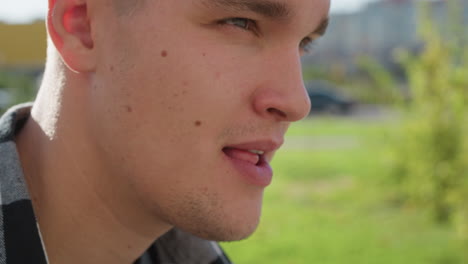 close up of young man licking lips subtly in bright outdoor setting with soft natural light highlighting facial texture and background of green field and distant buildings in smooth blur