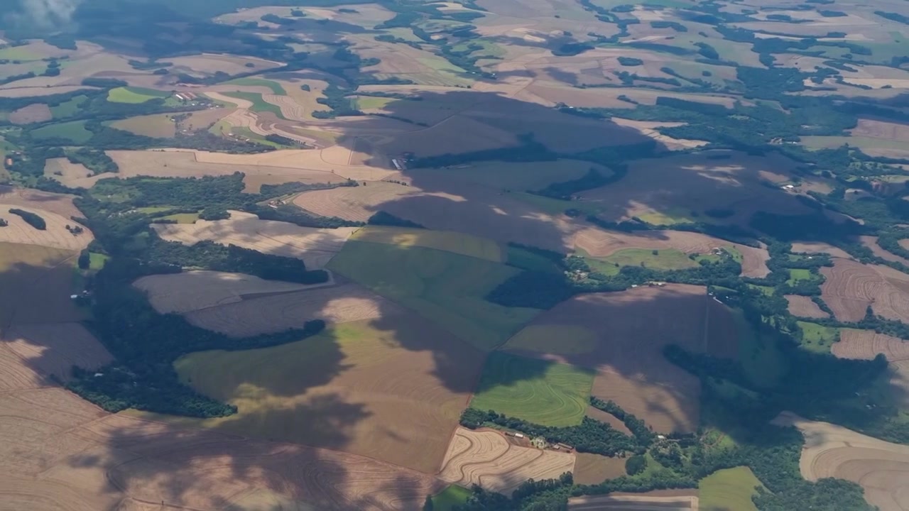 Letící skrz mraky nad Paraná, Brazílie, letecký pohled z okna odhaluje rozsáhlou zemědělskou půdu, venkovskou krajinu a barevné zemědělské vzory rozprostírající se po krajině dole