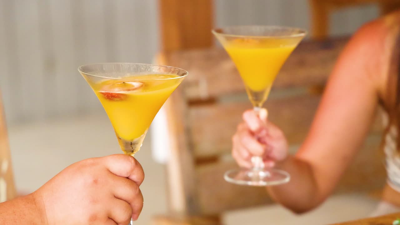 Two people clink glasses of vibrant yellow cocktails in a bright, relaxed outdoor environment with wooden furniture