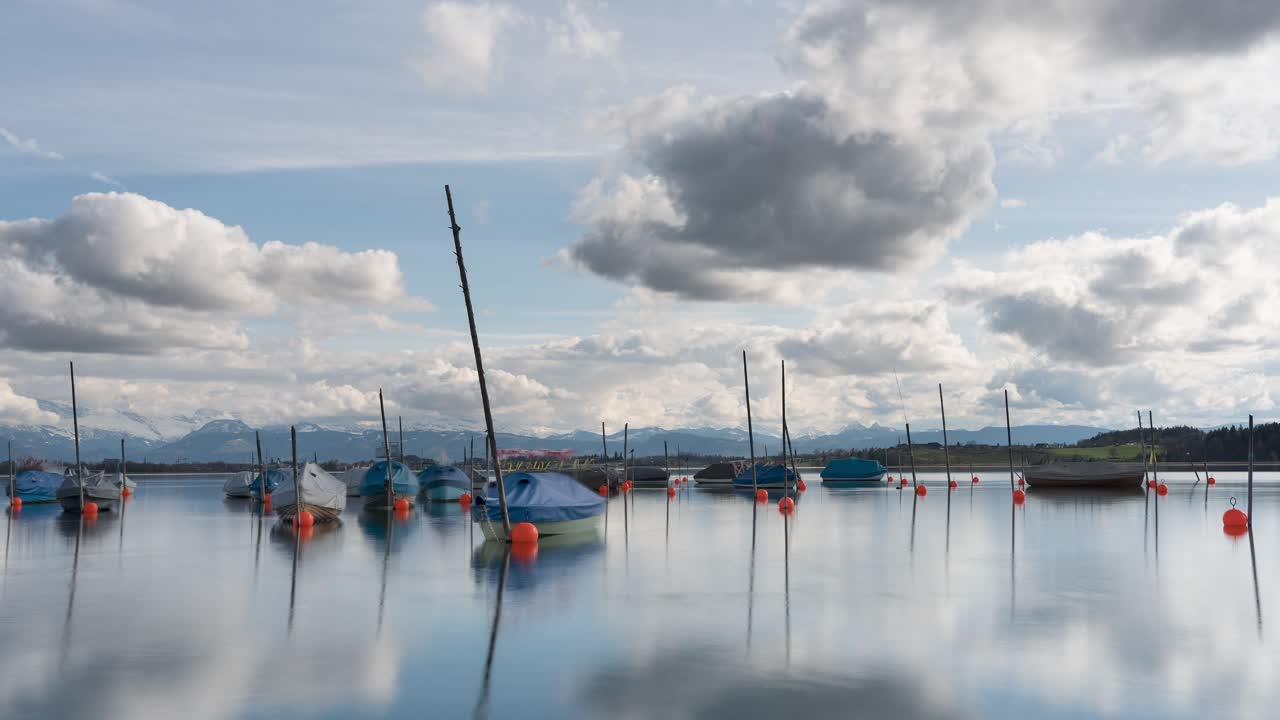 Spring arrives, and clouds drift over a natural landscape with a lake and fishing boats. The clouds form, and the sun shines. The water is calm. Time-lapse. In the background are the mountain ranges.
