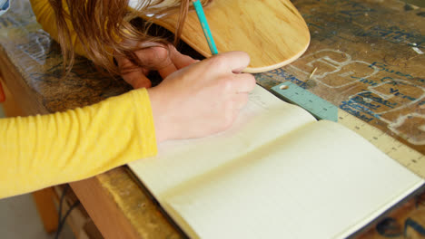 Close-up-of-young-blonde-woman-writing-in-diary-with-pencil-in-a-workshop-4k