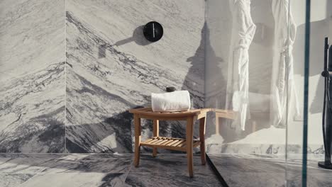 panning-shot-of-a-wooden-shower-stool-with-a-towel-and-rock-sitting-on-top-inside-of-a-luxury-shower-with-white-granite