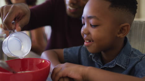 african american father smiling while pouring milk in cereal bowl on his son at home