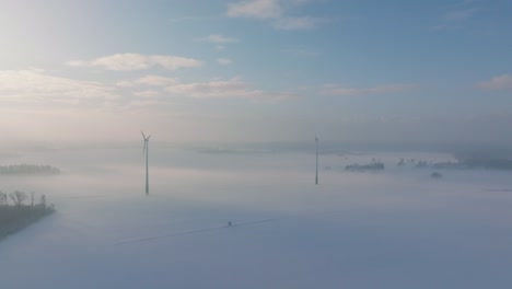 Establishing-aerial-view-of-wind-turbines-generating-renewable-energy-in-the-wind-farm,-snow-filled-countryside-landscape-with-fog,-sunny-winter-day,-wide-drone-shot-moving-forward