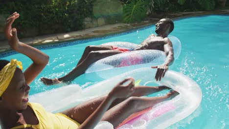 happy african american couple lying on inflatables in swimming pool splashing each other and smiling