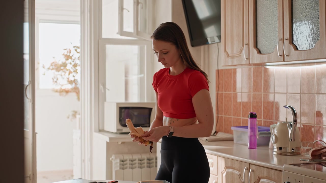 Junge Frau bereitet Banane in der Küche während der Morgenroutine zu, rotes Oberteil und schwarze Leggings, Wasserflasche und Mikrowelle auf der Arbeitsplatte, Läuferin bereitet Snack nach dem Lauf vor, warmes Sonnenlicht durchs Fenster, gemütlich