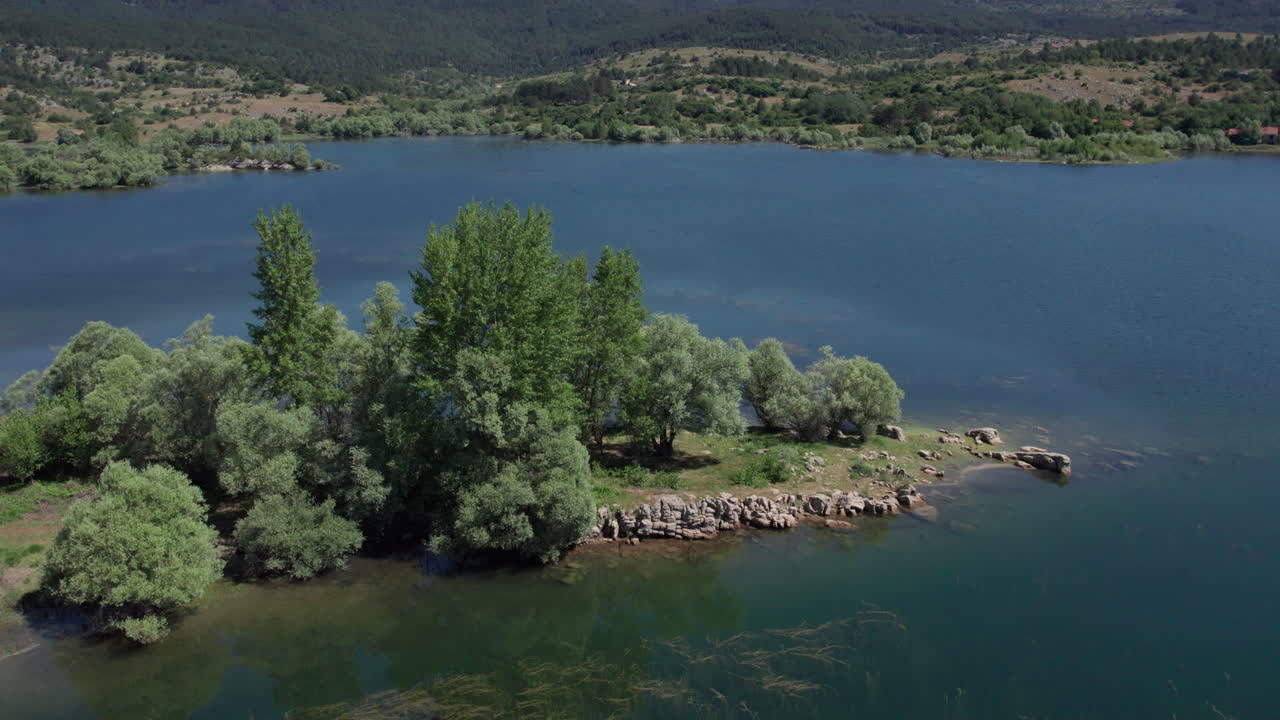 Letecký záběr dronem na zelený poloostrov zasahující do čisté modré vody jezera Tribistovo v Bosně a Hercegovině, obklopený letním lesem a klidnou krajinou, pro cestovatelská nebo přírodní videa.