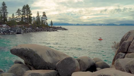 Large-rock-formations-with-a-beautiful-cold-blue-lake-surrounded-by-pine-trees-and-snow-capped-Sierra-Mountains-and-clouds---Sand-Harbor,-South-Lake-Tahoe,-Nevada---Wide-shot