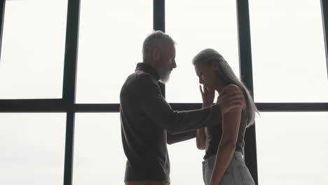 sad woman standing at window while her husband embracing and comforting her