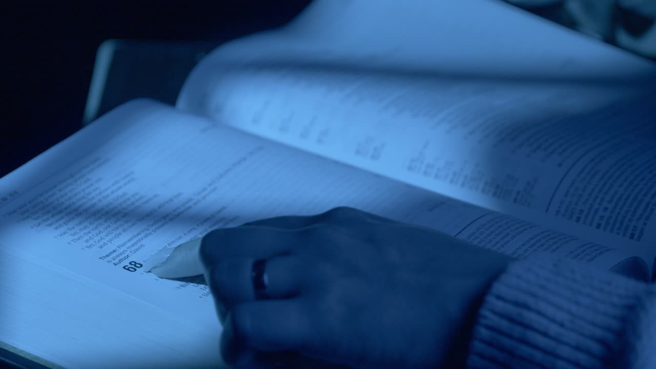 Uno scatto ravvicinato che cattura una mano che sfoglia delicatamente le pagine di una Bibbia sotto una luce blu soffusa. La messa a fuoco intima e l'ambiente tranquillo creano un'atmosfera riflessiva e serena.