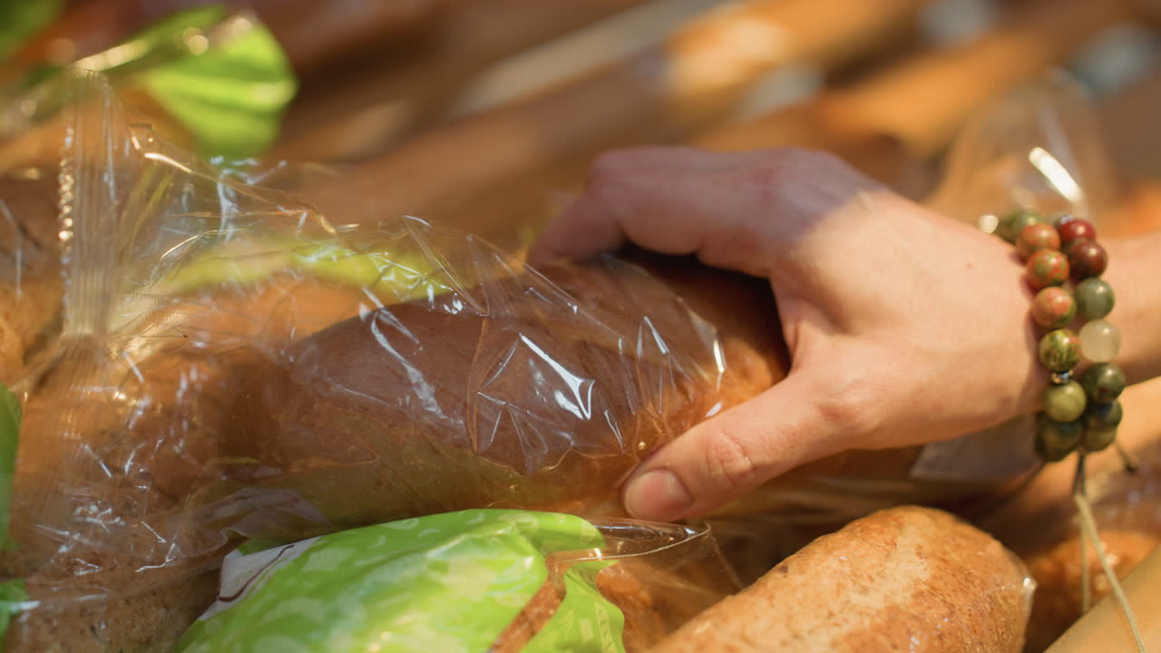 una mano con dos pulseras de cuentas tocando y recogiendo un pan en una tienda de comestibles. filmado con una cámara portátil, capturando la interacción detallada con el pan en la sección de panadería