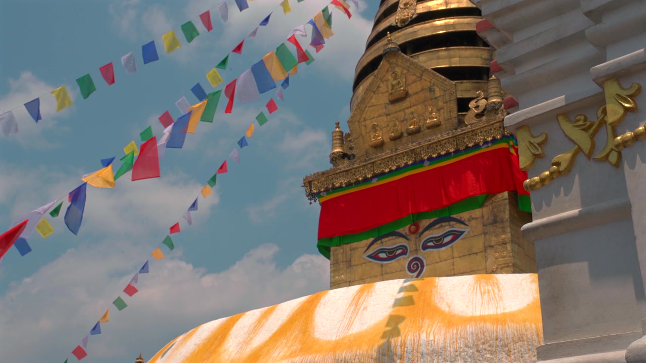 Una ripresa statica a media distanza e dal basso della guglia dorata e degli occhi della saggezza della Stupa di Swayambhunath, incorniciata da bandiere di preghiera colorate e una struttura di santuario bianca in Nepal.