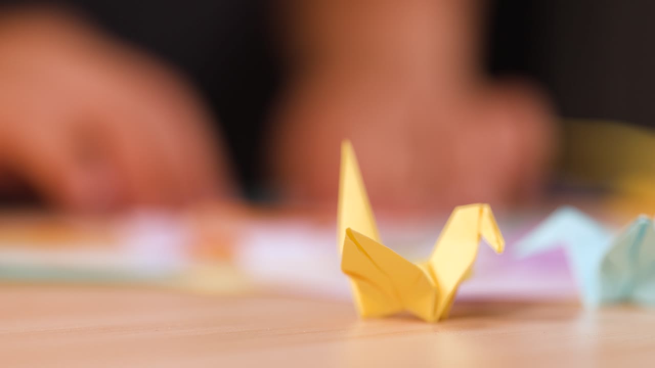 Primo piano di mani che piegano un foglio di carta, con una gru origami gialla completata a fuoco