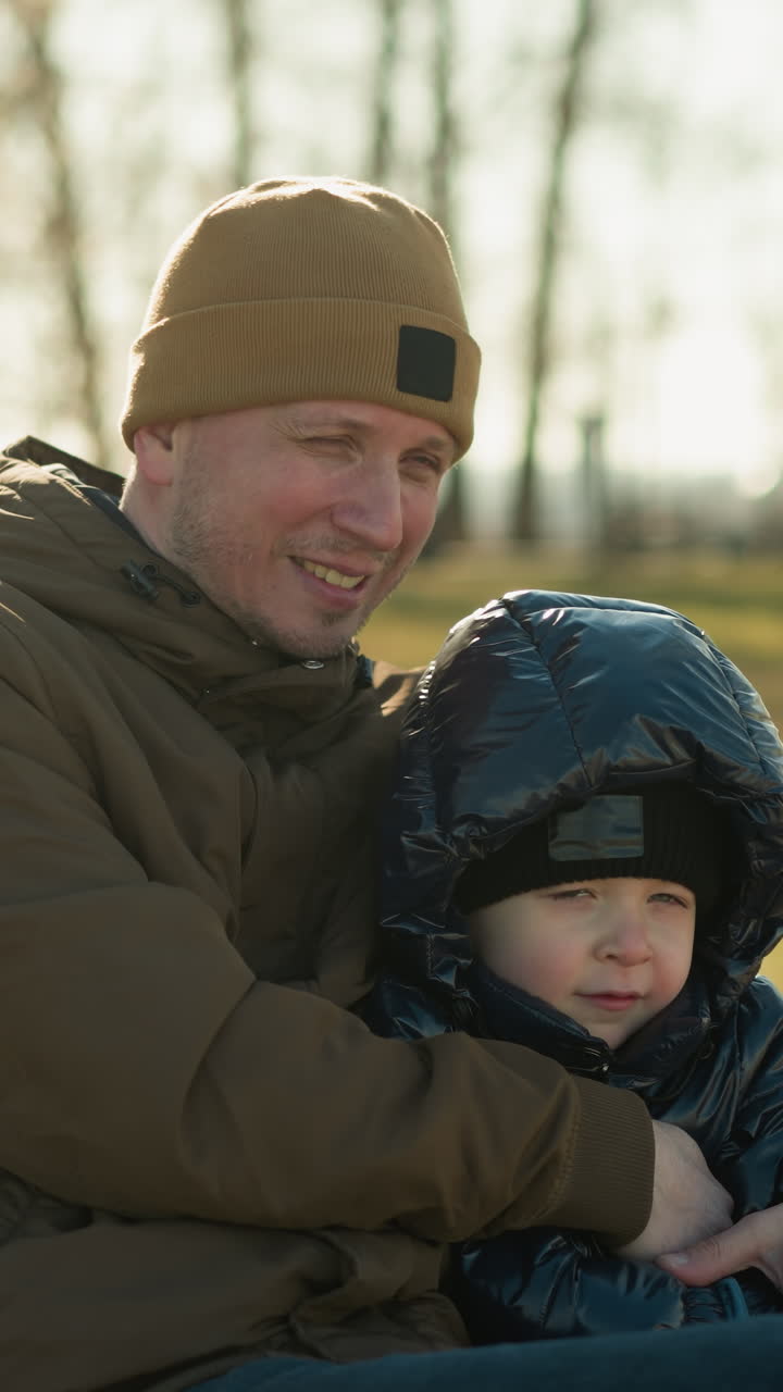 padre e hijo sentados juntos en un banco del parque, con el hijo en una chaqueta negra y el padre en una chaquette marrón, rodeados por el fondo borroso de árboles
