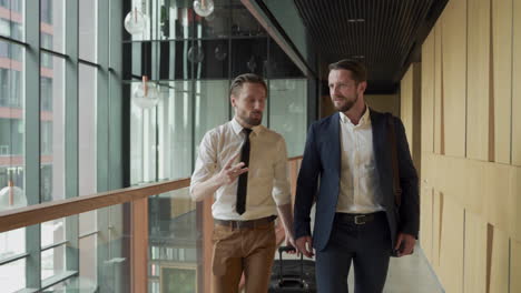 two young businessmen walking through the corridors of the airport carrying trolleys during a business trip