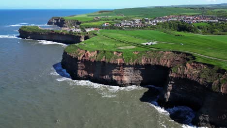 High-drone-view-of-north-yorkshire-coastline-showing-tall-cliffs-and-multiple-coves-and-textures-as-waves-crashing-from-the-ocean-into-the-cliffside-with-birds-flying-around