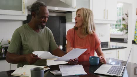 Happy-diverse-senior-couple-using-laptop-computer-and-paying-bills-in-kitchen