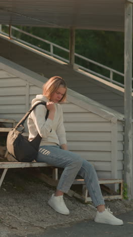 una mujer joven con una bolsa negra ajusta su posición de asiento en la tribuna oxidada del estadio, dando un paso adelante con la pierna derecha y reposicionando la bolsa