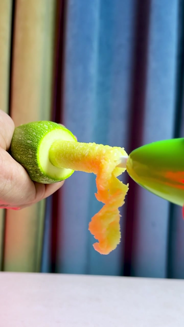 Un primer plano vertical de manos usando un descorazonador de plástico para vaciar un calabacín fresco. La preparación de alimentos saludables se realiza en interiores sobre una mesa, con un fondo borroso de rayas de colores.