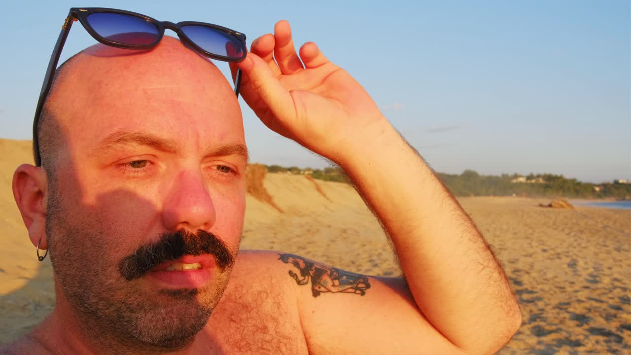 caucásico calvo de cabeza afeitada hombre con bigote y gafas de sol en la playa durante la puesta del sol