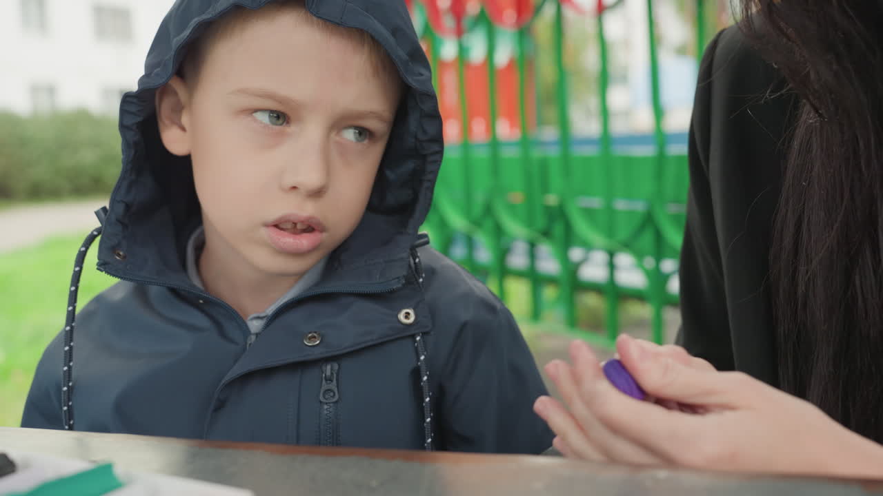 Ragazzo bianco con giacca con cappuccio a cui viene offerta una piccola gomma viola mentre si prepara per il ritorno a scuola, mano attenta del genitore nell'inquadratura, tavolo da gioco all'aperto, espressione curiosa che si trasforma in accettazione spontanea.