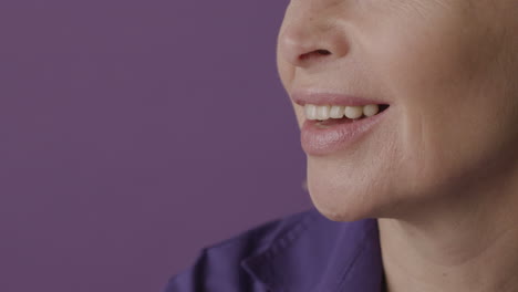 close up view of half face of blonde mature woman with purple shirt smiling on purple background