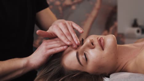 close-up portrait of male massage therapist's hands giving gentle facial massage to young woman in a massage room with soft lighting