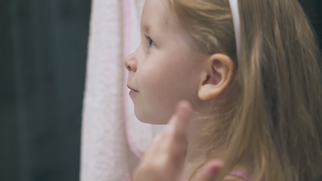 긴 머리카락을 가진 행복한 소녀는 밝은 욕실에서 거울을 본다