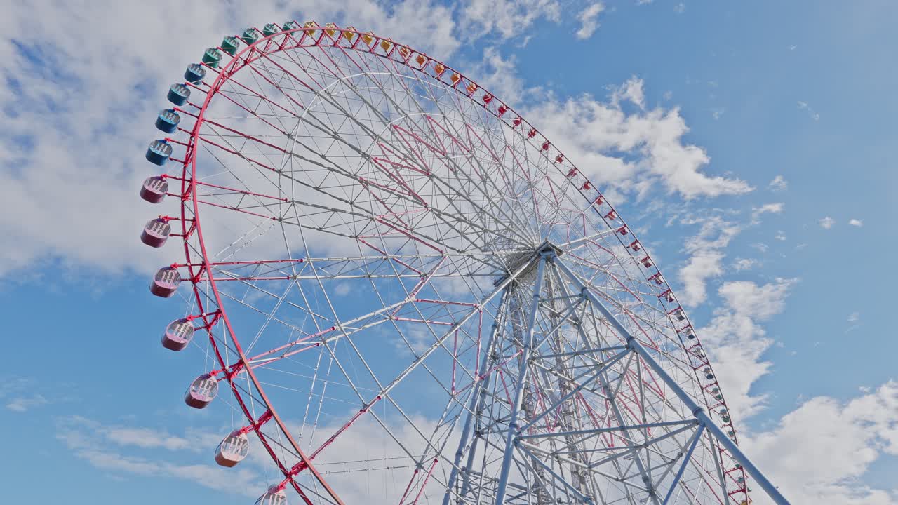 Wide low-angle view showing the entire massive white and red Ferris wheel turning slowly against a bright blue sky dotted with clouds