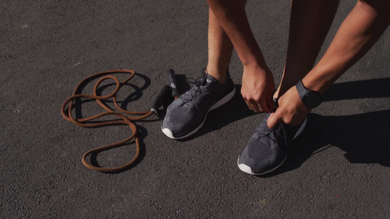 Low section of african american man exercising in city with skipping rope, tying shoelaces in street