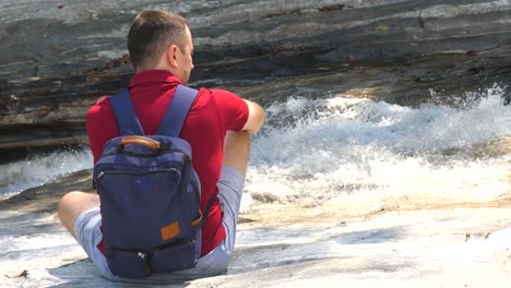 solo viajero masculino con una mochila se sienta cerca de un río de montaña burbujeante