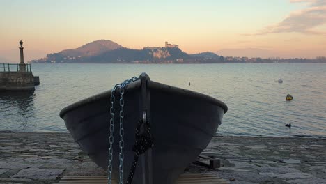 Fischerboot-Aus-Dem-Wasser-Und-Mit-Einer-Eisenkette-Festgebunden,-Mit-Blick-Auf-Den-Lago-Maggiore-Und-Die-Burg-Von-Angera,-Statisch