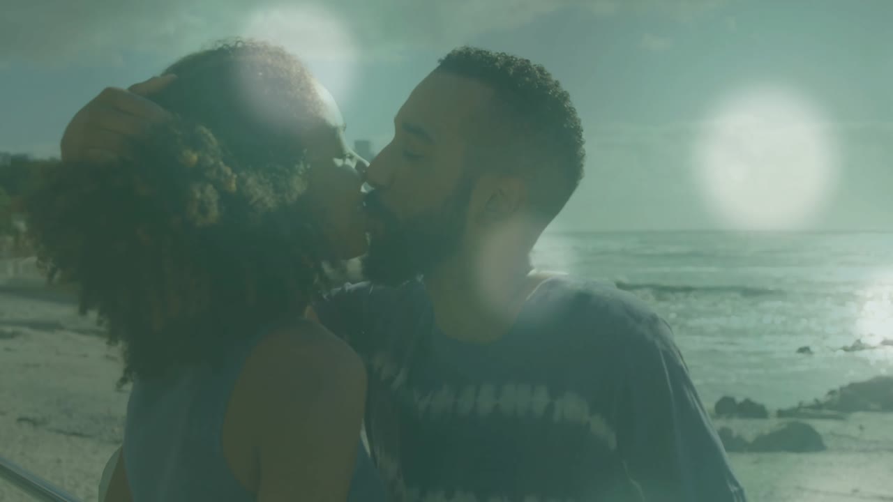 Coppia che si scambia un bacio in spiaggia dopo che l'uomo sistema una molletta rossa per capelli, con bagliori dell'obiettivo fluttuanti per scopi di marketing.