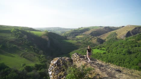 Slow-mo-wide-shot-of-woman-walking-towards-the-edge-of-a-cliff-above-Thor's-cave,-Ashbourne,-Peak-District,-England