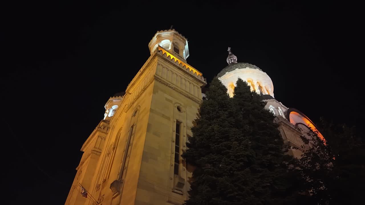 Walking around the illuminated Metropolitan Cathedral of the Assumption in Cluj-Napoca, Romania, showing its domes and towers glowing warmly against the night sky